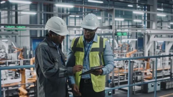 多种族经理和非裔美国人汽车工厂工程师穿着制服，使用平板电脑。汽车行业4.0制造员工讨论车辆装配厂的工