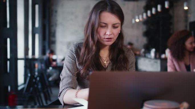 年轻快乐迷人体贴的高加索黑发女性创意作家用笔记本电脑在咖啡馆工作做笔记。