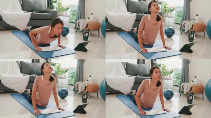 年轻迷人的亚洲女性穿着运动服，在平板电脑上观看健身在线视频，为初学者在家里的客厅练习瑜伽。