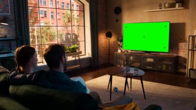 真正的情侣在家里度过时光，坐在沙发上，在他们时尚的阁楼公寓里看绿屏模拟显示的电视。男人和女人流媒体电