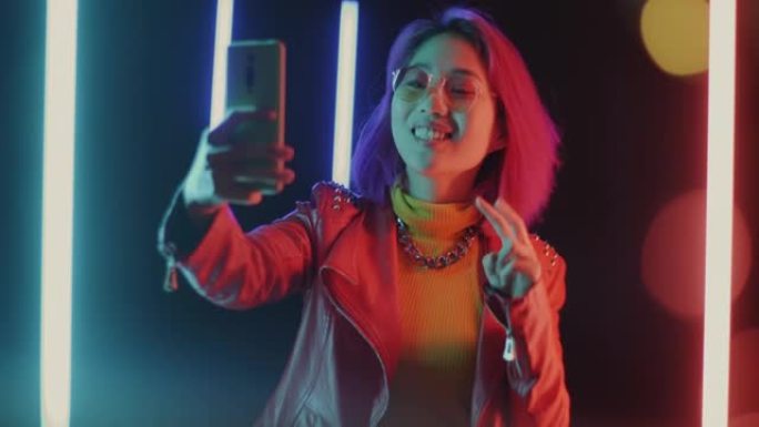 在未来霓虹灯背景下为智能手机相机拍照的快乐亚洲学生肖像