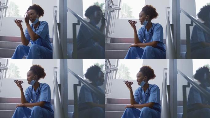 混血女医生戴着口罩坐在医院的楼梯上讲智能手机