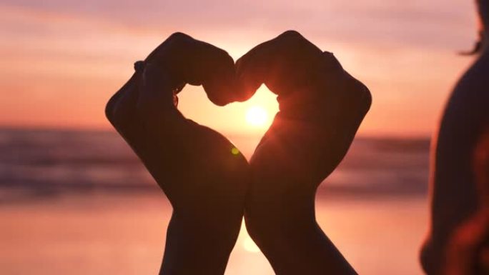 女人在海滩上的爱，心和手，以争取自由，支持和幸福，在日落时签名。剪影，自然和对女孩的尊重，以浪漫，灵