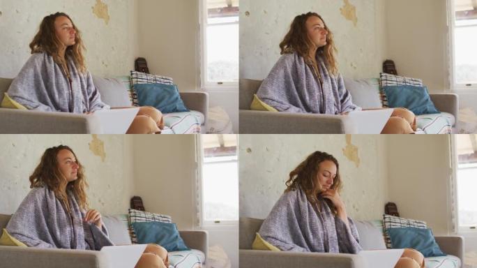 体贴的白人妇女穿着毯子坐在沙发上，用笔记本电脑在阳光明媚的小屋客厅