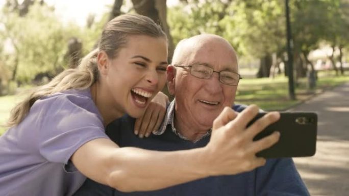 一个快乐的年轻照顾者，在外面的公园里用手机自拍时，和她的老人一起玩得很开心。中风后，坐在轮椅上的老人