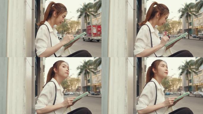 在城市现代城市中独自一人坐在户外时，穿着时尚办公服装的成功年轻亚洲女商人使用数字平板电脑和打字短信。