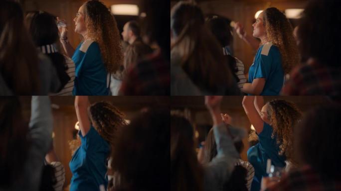 一位穿着蓝色t恤的年轻女性的肖像站在酒吧里的一群体育迷中，看着电视转播。球迷庆祝球队进球并赢得冠军。