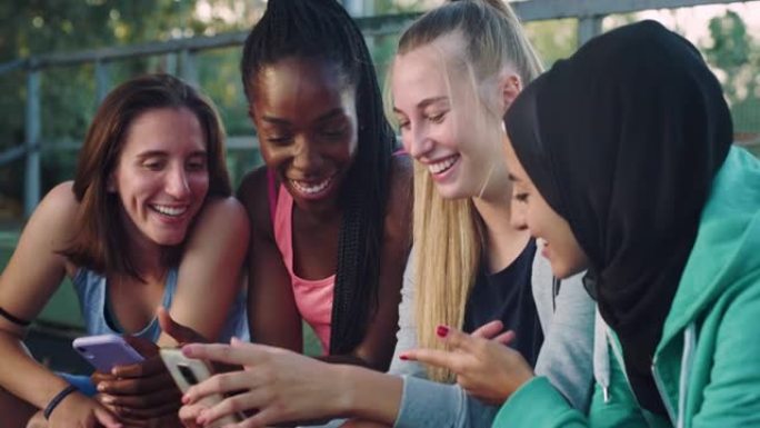 一群年轻的多种族女性青少年坐在公园里，笑着指着智能手机。一群朋友一起使用技术重温童年记忆和照片