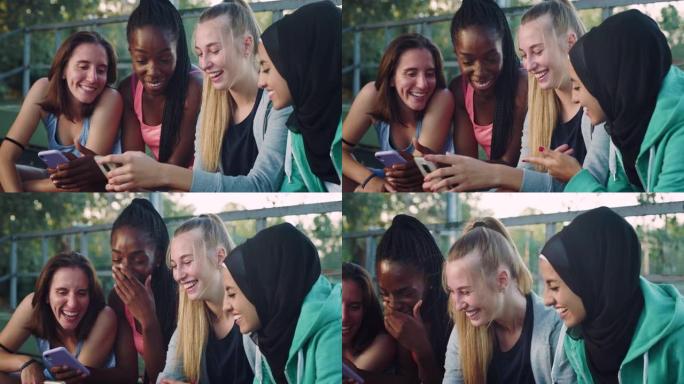 一群年轻的多种族女性青少年坐在公园里，笑着指着智能手机。一群朋友一起使用技术重温童年记忆和照片