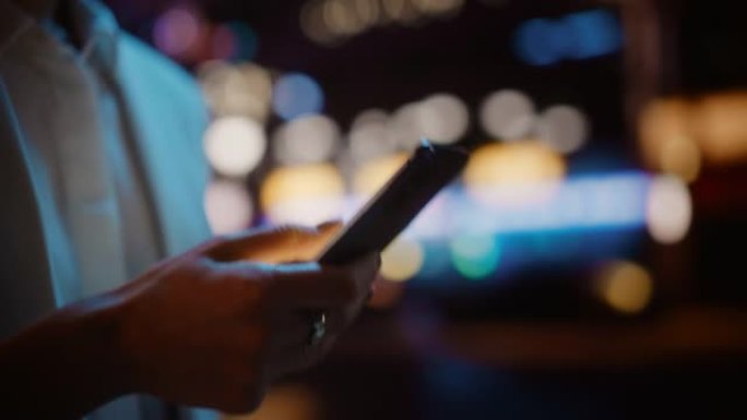 使用智能手机在充满霓虹灯的夜晚城市街道上行走，专注于女性手。女人拿着手机，发布社交媒体，在线购物，发