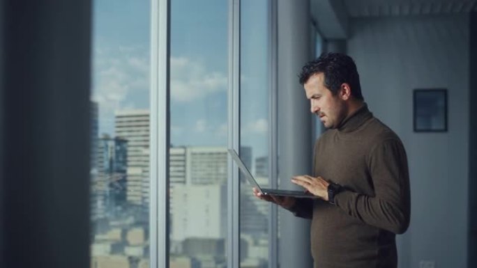 体贴的中东商人站在大城市窗户附近的办公室时使用笔记本电脑。成功的数字企业家从事电子商务投资的数据分析