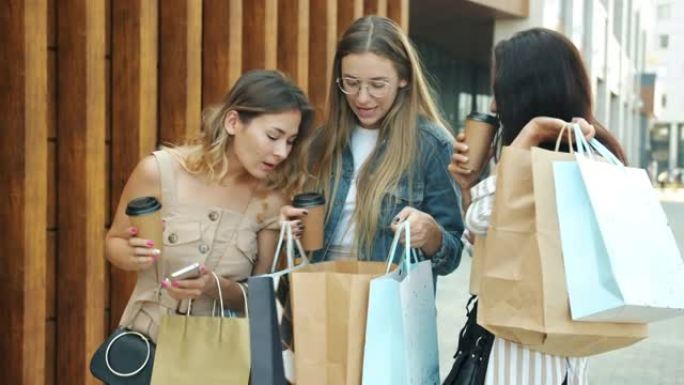 开朗的年轻女性朋友在户外的购物袋中碰碰咖啡杯和聊天，看着购物