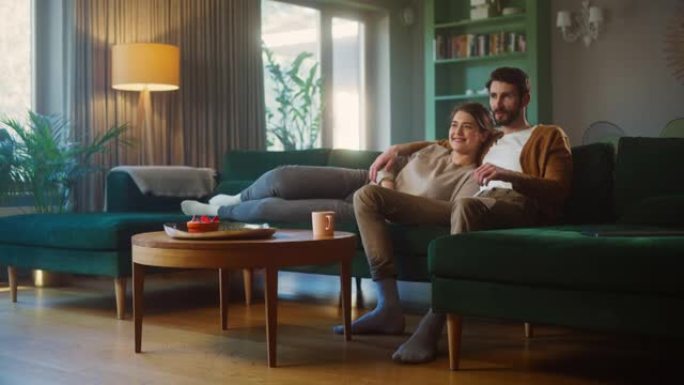 夫妇坐在客厅的沙发上看电视。女朋友和男朋友躺在一起拥抱，用遥控器切换频道。在家里度过周末，穿着时尚的