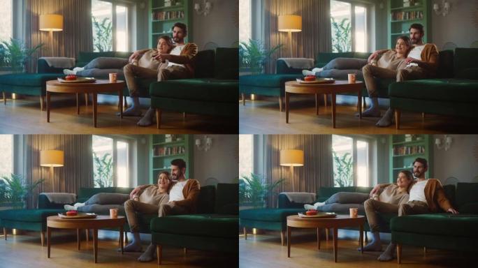 夫妇坐在客厅的沙发上看电视。女朋友和男朋友躺在一起拥抱，用遥控器切换频道。在家里度过周末，穿着时尚的