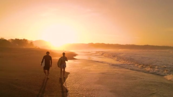 空中轮廓: 两名冲浪者在海滩上行走，日出时冲浪