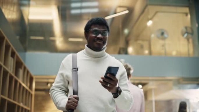 年轻黑人男子的肖像在繁忙的办公室走廊里微笑着检查手机时感到自信。他正在用智能手机浏览互联网。低角度慢