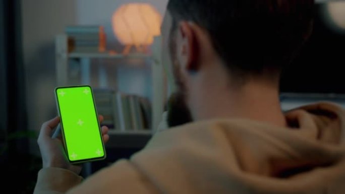 年轻人的肩膀上的镜头在晚上在家室内看着绿色色度键智能手机屏幕