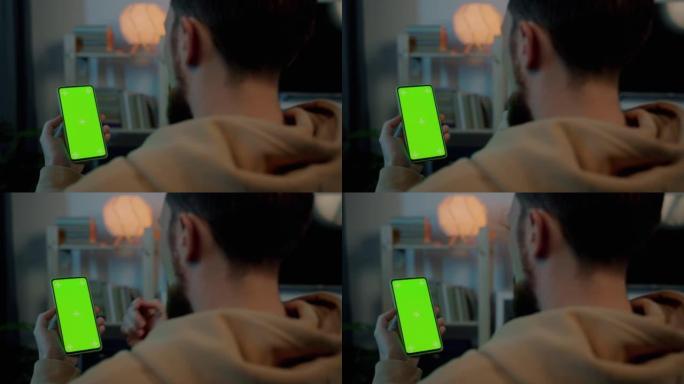 年轻人的肩膀上的镜头在晚上在家室内看着绿色色度键智能手机屏幕