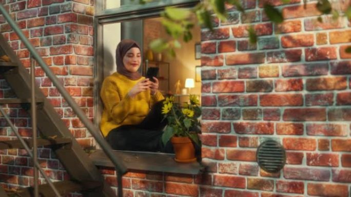 年轻的穆斯林女性坐在多层砖房的窗台上时使用智能手机。在社交媒体上与家人和朋友联系时，富有创造力的女性