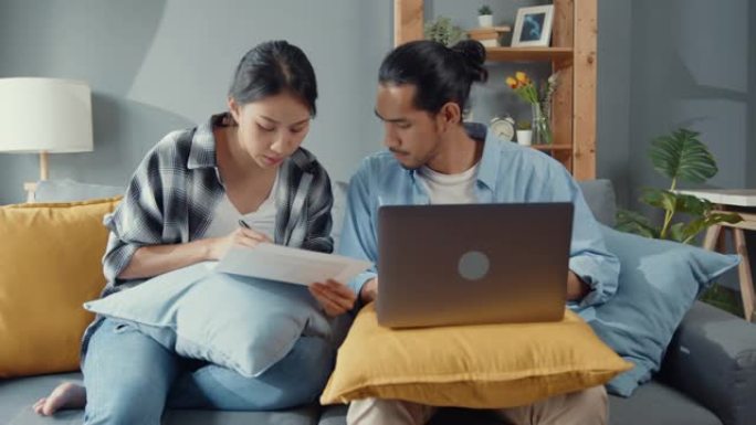 亚洲年轻夫妇坐在沙发上认真关注笔记本电脑检查文件纸质支付账单在线计划客厅预算费用。