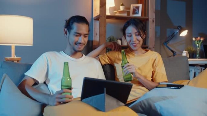 快乐的年轻亚洲夫妇享受夜间派对活动在线坐沙发使用平板电脑视频通话与朋友在家里的客厅通过视频通话吐司喝