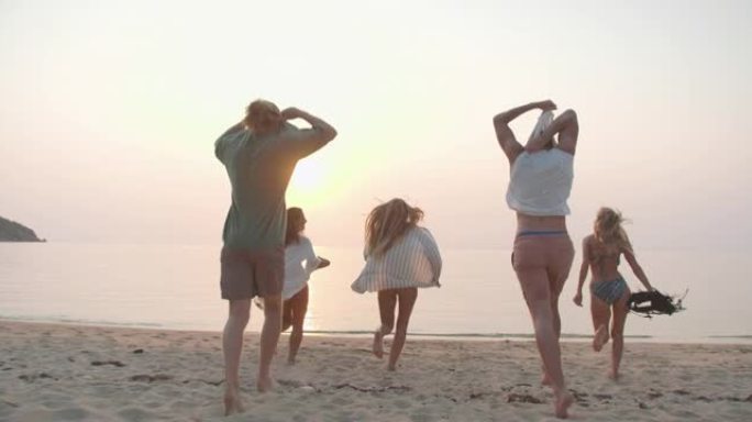 后视图，很高兴一群年轻的多民族的五个朋友一起奔跑，脱下衣服，一起在沙滩上沿着日落海的边缘游泳，玩得开