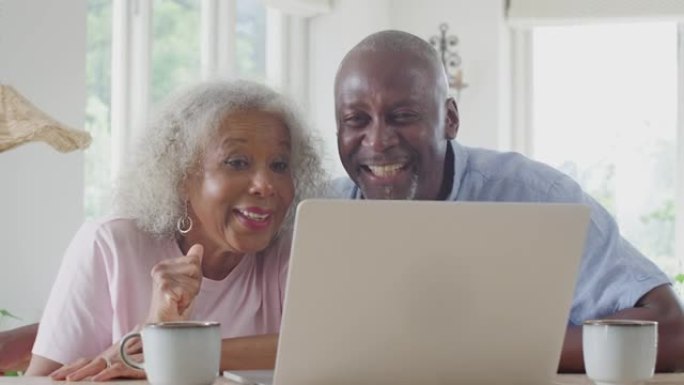 高级夫妇坐在家里的桌子旁，用笔记本电脑进行视频通话