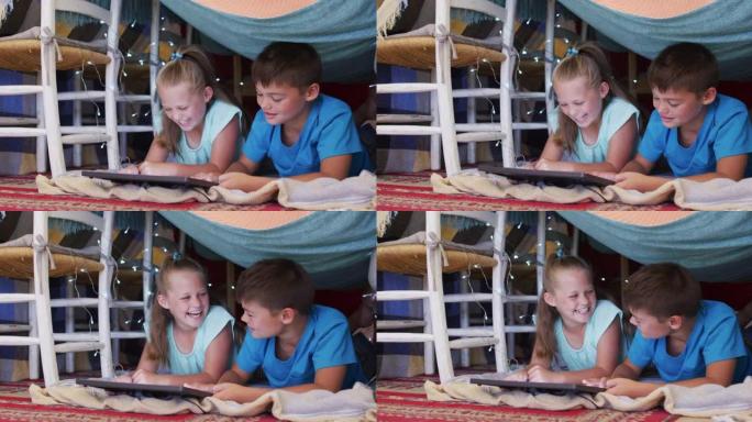 高加索兄弟姐妹在家里的毯子下使用数字平板电脑时微笑着