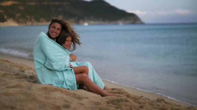 一个年轻无忧无虑的快乐微笑的母亲和被拥抱在条纹封面下的小女儿的真实照片在日落时在海边享受着他们的时光