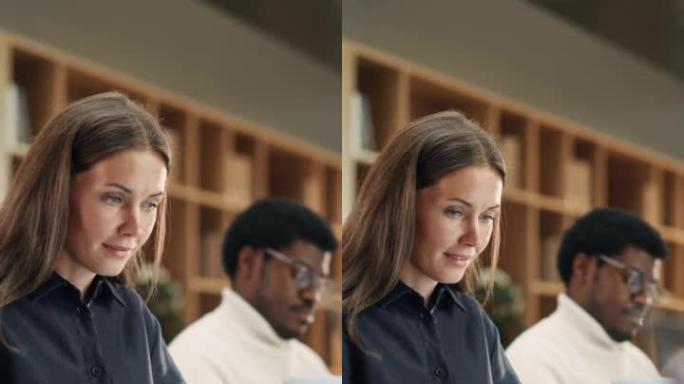 垂直镜头: 使用笔记本电脑在多元化办公室工作的白人妇女的肖像。女顾问微笑着回复电子邮件。在后台工作的