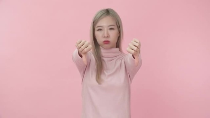 穿着粉红色长袖t恤的年轻美丽的亚洲女人的肖像，在粉红色背景下的相机上露出拇指朝下的不喜欢的表情。情感