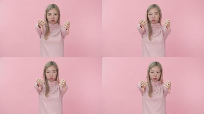 穿着粉红色长袖t恤的年轻美丽的亚洲女人的肖像，在粉红色背景下的相机上露出拇指朝下的不喜欢的表情。情感