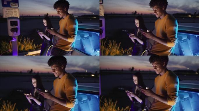 SLO MO年轻夫妇在晚上为电动汽车充电时使用智能手机
