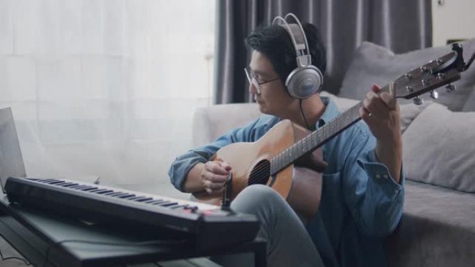中等镜头亚洲成年男性在家中客厅弹奏键盘钢琴和原声吉他。弹吉他创作音乐或在笔记本电脑上录制音乐的人。音