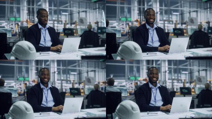 一位快乐的年轻非洲裔美国工程师在汽车装配厂的办公室里在笔记本电脑上工作的肖像。在技术开发设施中从事车