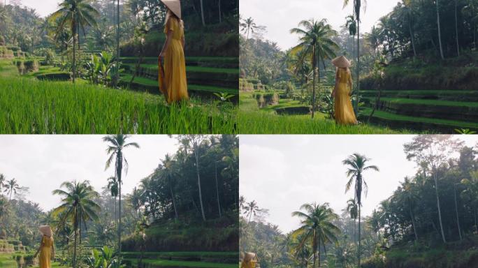 女人穿着黄色连衣裙，戴着圆锥形帽子，在文化景观中探索郁郁葱葱的绿色大米露台，穿越巴厘岛印度尼西亚发现