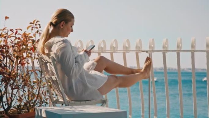 金发碧眼的女人坐在酒店房间的阳台上，可以看到大海，低头看着她的智能手机，双腿伸出