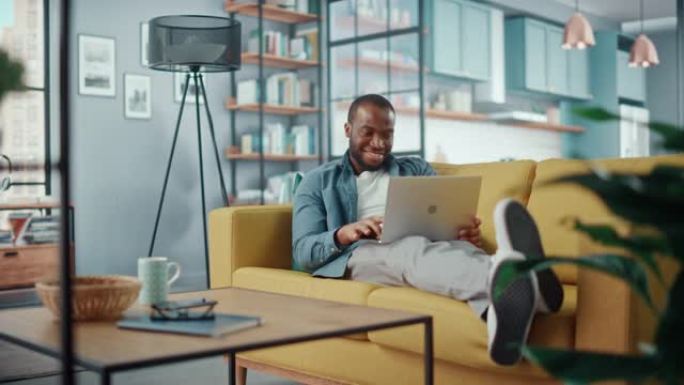 英俊的黑人非洲裔美国男子躺在舒适的客厅的沙发上，在笔记本电脑上工作。自由职业者在家工作。浏览互联网，
