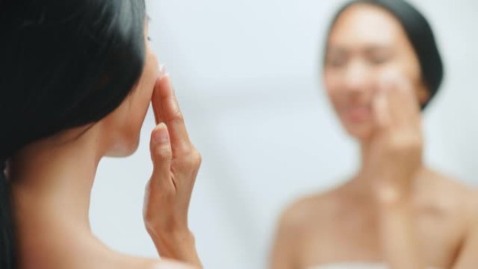 美丽的亚洲女人的肖像轻轻地涂抹面霜面膜，用性感的触摸，看着浴室的镜子。年轻女性用天然化妆品护肤产品使