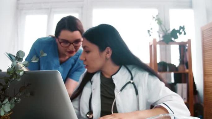 年轻的多民族药房医生在笔记本电脑网络摄像头上咨询远程客户，与穿着蓝色制服的助手交谈。