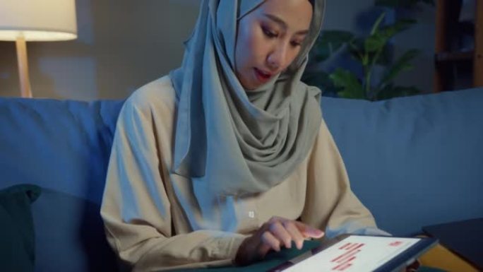 迷人开朗的年轻亚洲穆斯林美女戴头巾随意坐在沙发上，配有笔记本电脑，晚上在家里舒适的客厅使用数字平板电