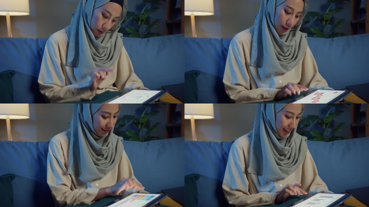迷人开朗的年轻亚洲穆斯林美女戴头巾随意坐在沙发上，配有笔记本电脑，晚上在家里舒适的客厅使用数字平板电