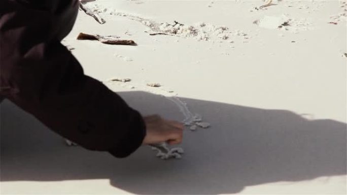 十几岁的女孩穿着夹克，在海滩上的白色沙滩上画她的名字。特写。