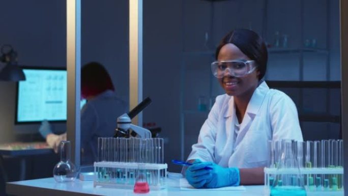 一位年轻的迷人的非裔美国女科学家戴着防护眼镜，微笑着坐在相机上，坐在她工作场所的现代实验室中