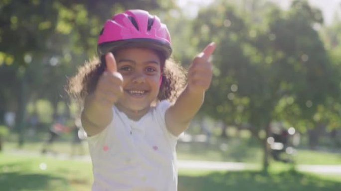 在公园阳光明媚的日子里，快乐的兴奋的小女孩戴着粉红色的头盔，一边跳舞，一边竖起大拇指。可爱的孩子积极