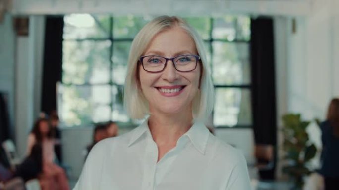 快乐的高级老师，教育专业的女商人在办公室慢动作时戴上眼镜对着镜头微笑。