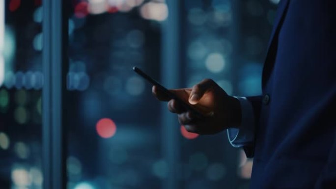 夜间办公室: 成功的黑人商人穿着西装站立，使用智能手机。CEO浏览互联网，管理电子商务软件中的社交媒