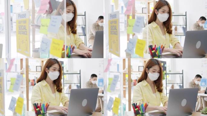 亚洲女性在新的正常情况下戴口罩，防止病毒，同时使用笔记本电脑，办公室用丙烯酸隔板隔开。