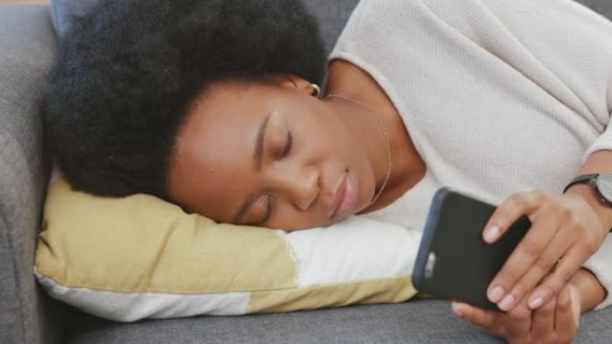 疲惫的黑人妇女在家休息时在电话上发短信。精疲力竭的非洲妇女在沙发上浏览社交媒体时入睡，利用空闲时间小