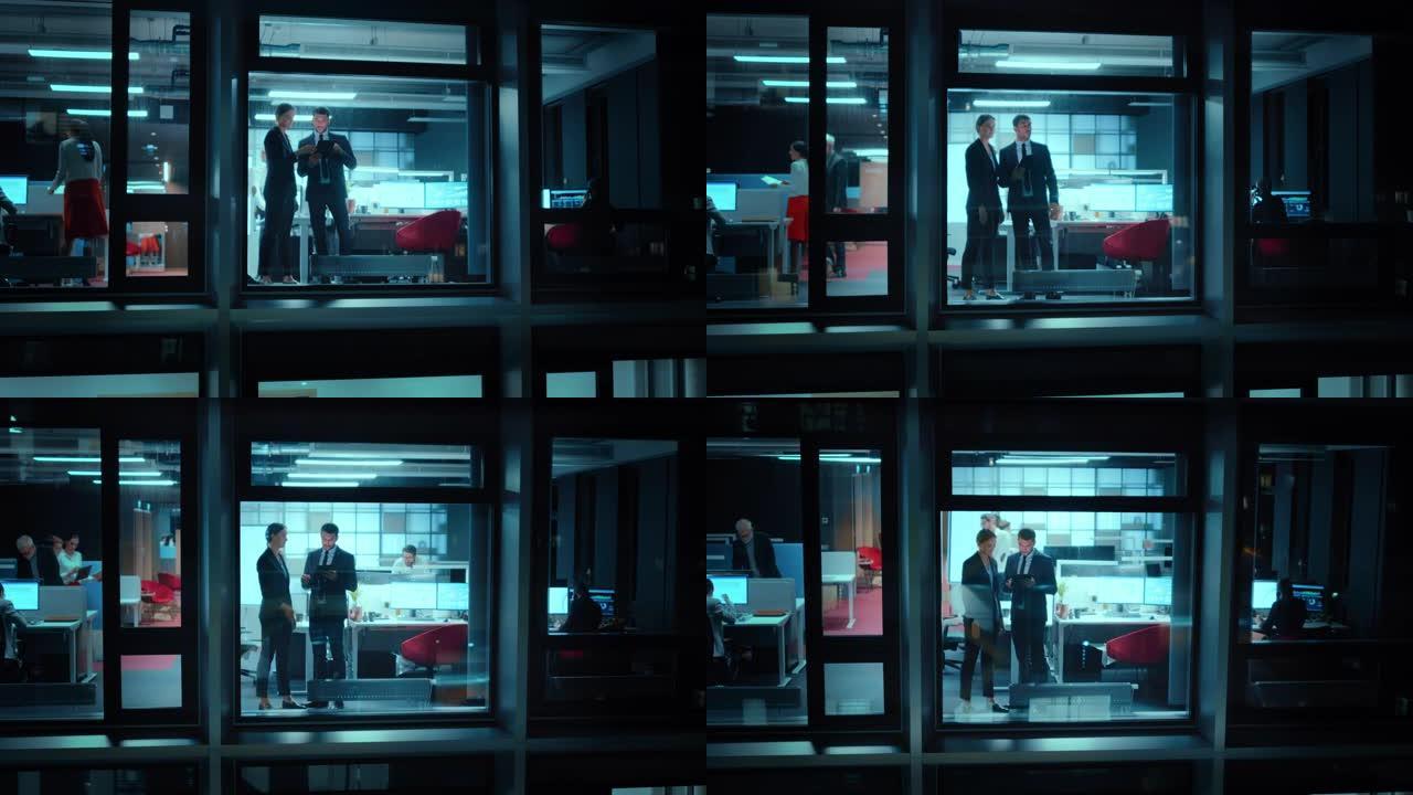 外面的镜头: 两个商业伙伴在办公室窗户旁边使用平板电脑。员工谈财务战略与业务发展同事在他们身后工作。